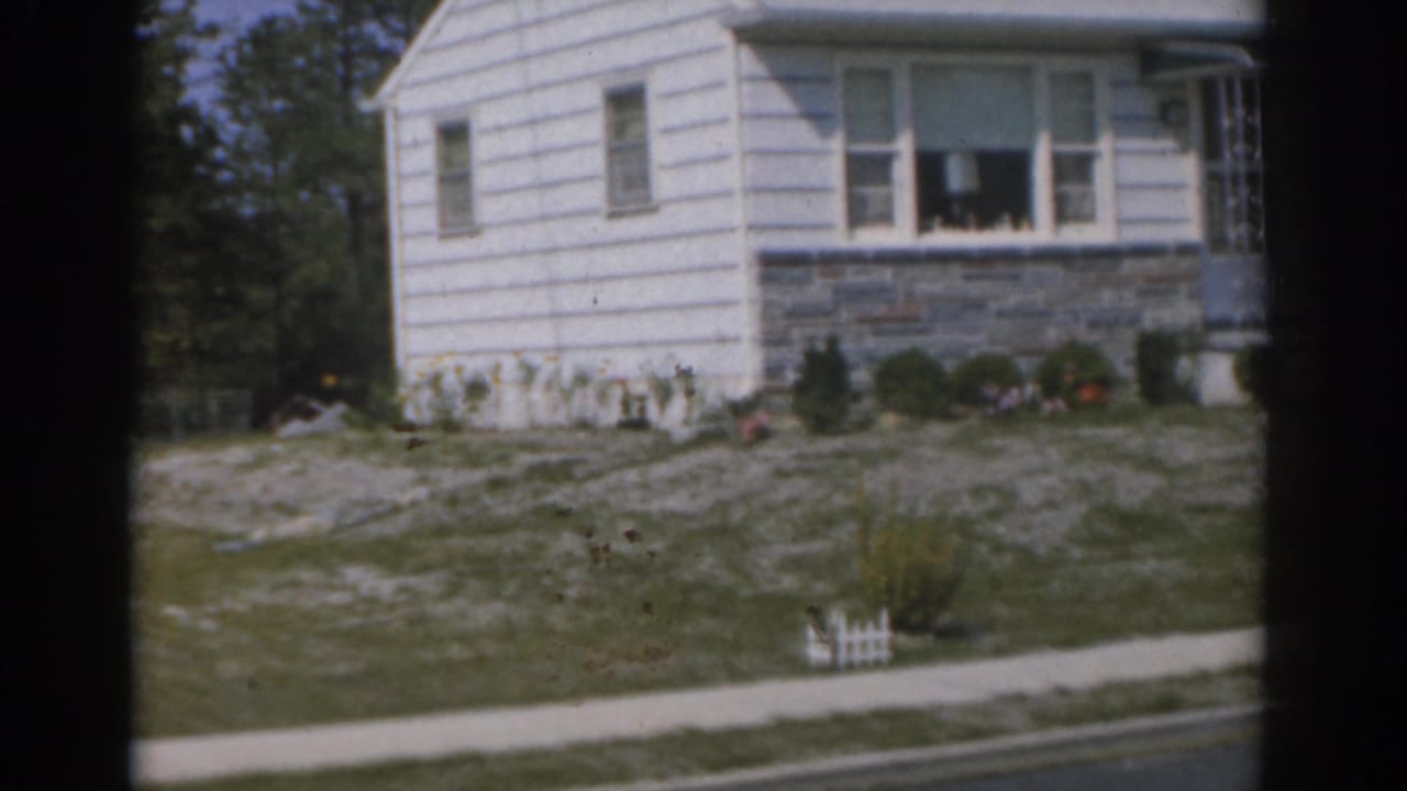 1963 New Jersey 8mm frame showing a modest white house exterior with grey stone wall, shrubs, sidewalk, and small white garden fence.