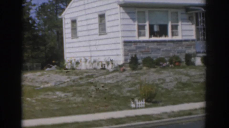 1963 New Jersey 8mm frame showing a modest white house exterior with grey stone wall, shrubs, sidewalk, and small white garden fence.