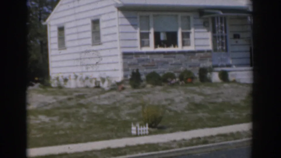 1963 New Jersey home-movie frame of a white paneled house front with picture window, grey stone accent wall, shrubs, and sidewalk.