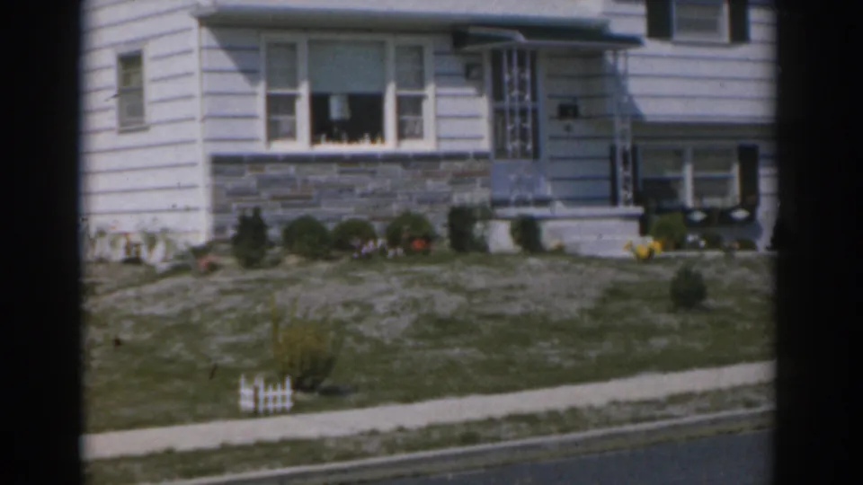 1963 New Jersey home-movie frame showing a white suburban house exterior with entry steps, picture window, stone wall, shrubs, and lawn.