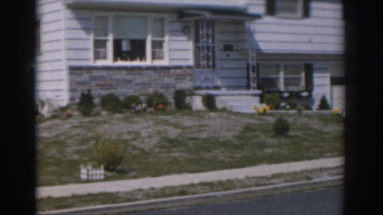 1963 New Jersey home-movie frame showing a white-sided suburban house with stone lower wall, porch steps, lawn, and sidewalk.
