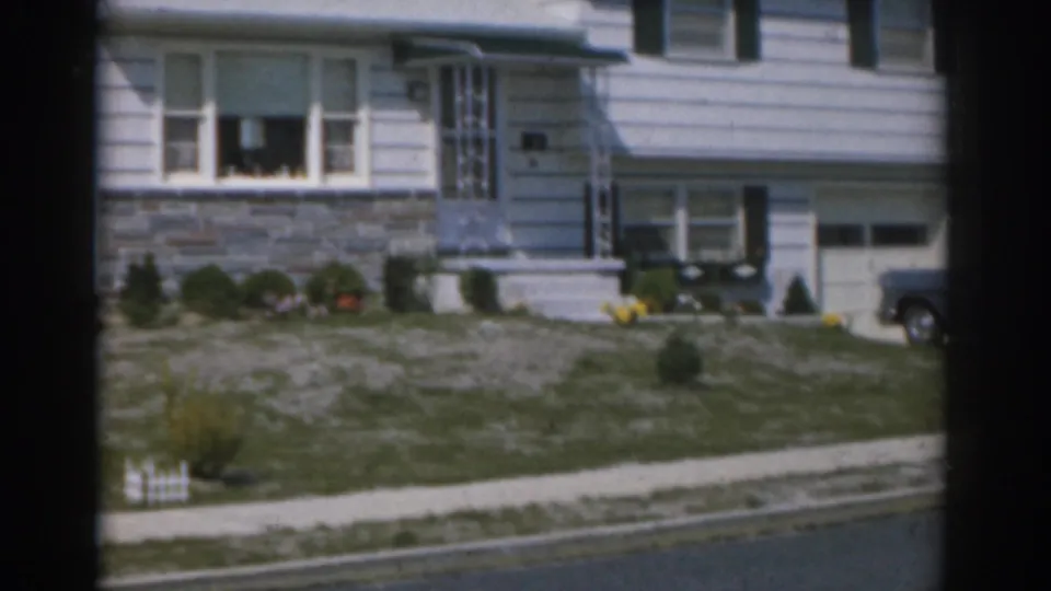 1963 New Jersey residential exterior with a white house, landscaped front lawn, sidewalk, and a blue car beginning to enter frame.