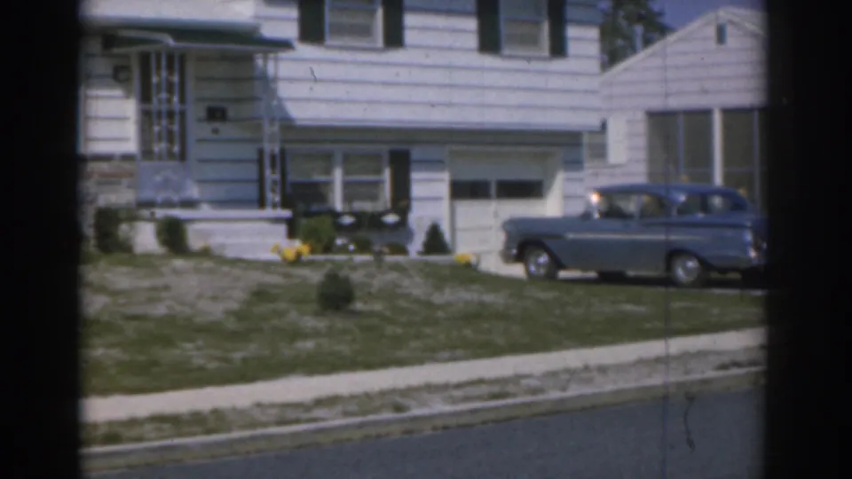 1963 New Jersey residential scene with a blue station wagon stopped in the driveway next to a white-sided suburban house.