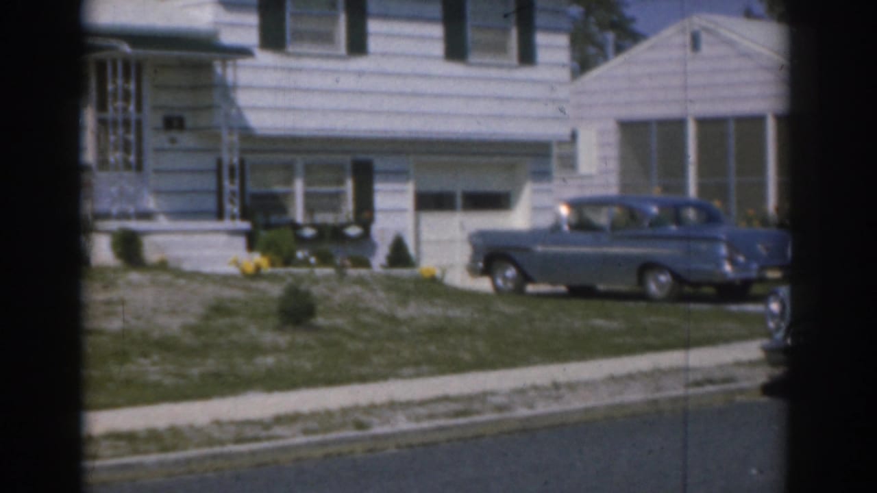 1963 New Jersey home-movie frame of a blue car parked at a white-sided suburban house with lawn and street visible.