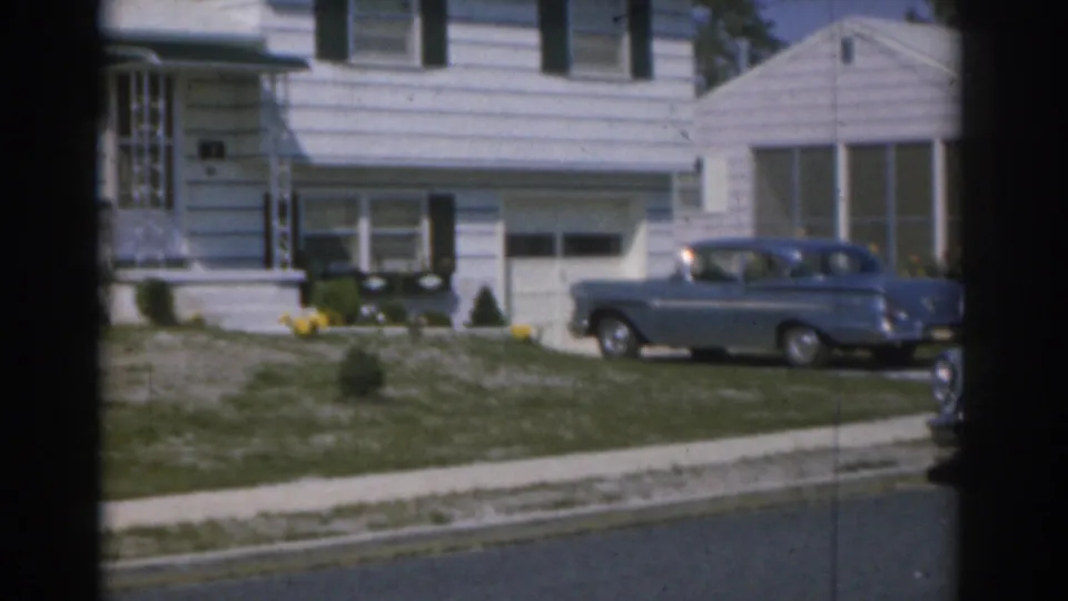 1963 New Jersey home-movie frame of a blue car parked at a white-sided suburban house with lawn and street visible.