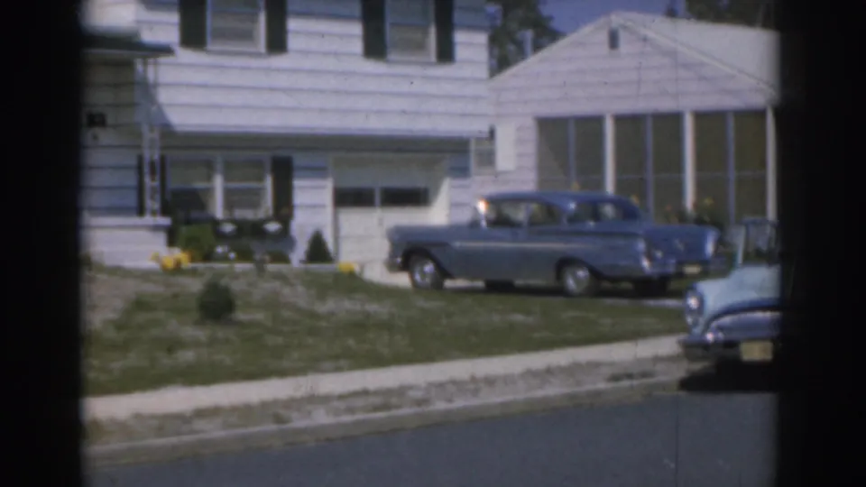 1963 New Jersey suburban street view with a blue parked car, white house, lawn, and the front of another car at frame edge.