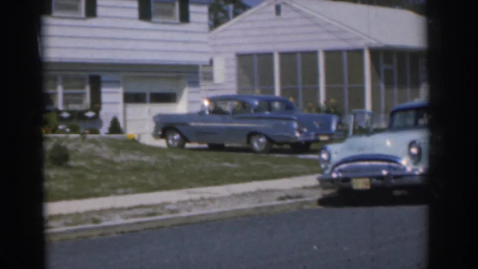 1963 New Jersey home-movie frame showing two blue cars near a white suburban house, with lawn, curb, and street in view.