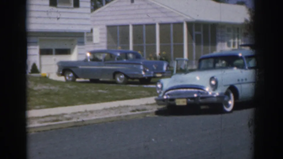 1963 New Jersey residential exterior with a parked blue car in the driveway and another blue car prominent on the street.