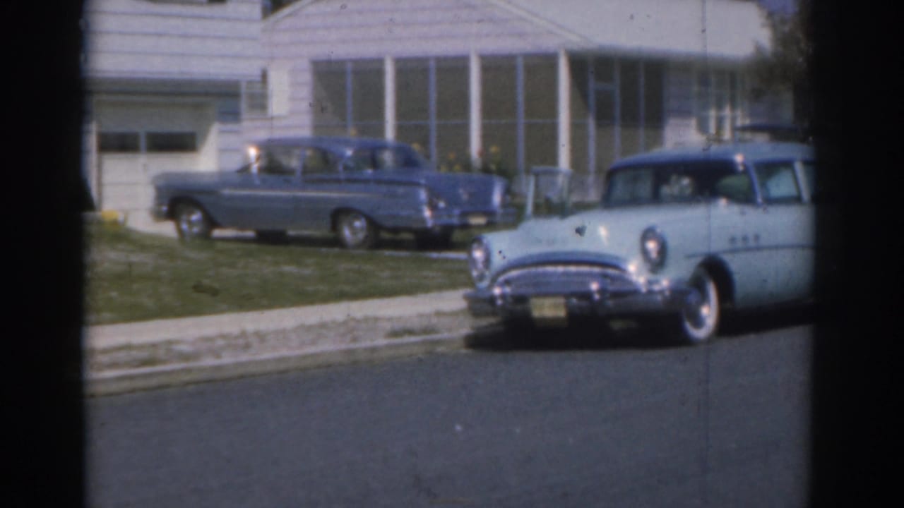 1963 New Jersey 8mm frame of a light blue sedan on a residential street near a parked blue car and white-sided houses.