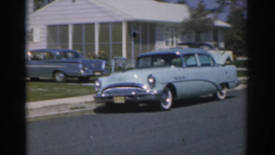 1963 New Jersey suburban street view with a light blue sedan, parked blue car, green lawns, and white residential houses.