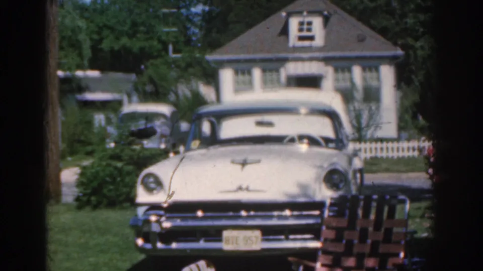 1963 New Jersey home-movie frame showing a cream-colored parked car before a house, lawn, shrubs, and white picket fence.