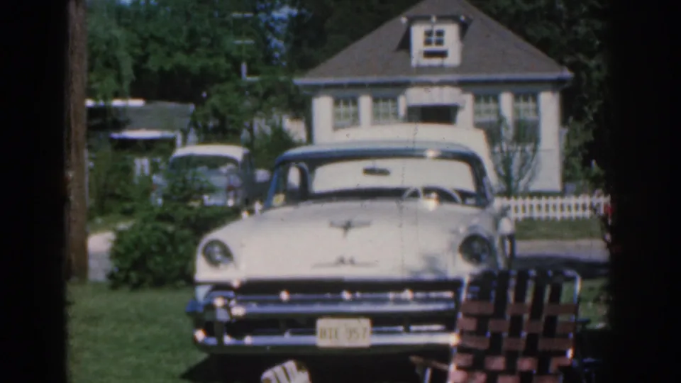 1963 New Jersey residential scene with a cream-colored parked car, white picket fence, house facade, lawn, and trees.