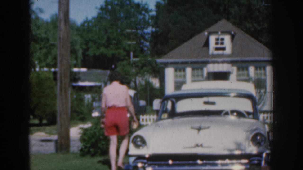 1963 New Jersey home-movie frame showing a woman walking near a white vintage car, with a house, lawn, trees, and fence behind her.
