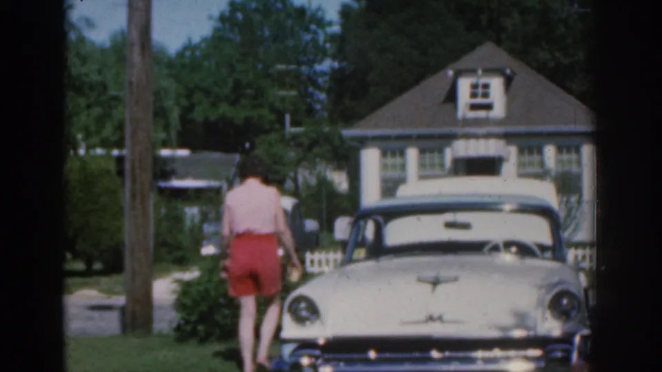 1963 New Jersey home-movie frame showing a woman walking near a white vintage car, with a house, lawn, trees, and fence behind her.