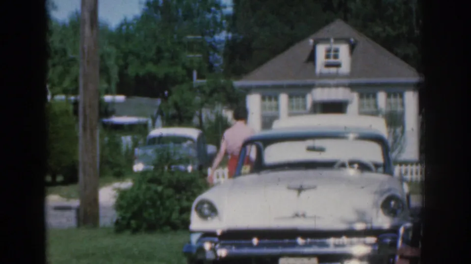 1963 New Jersey frame with a woman moving across a residential yard near a white vintage car, framed by a house, driveway, trees, and fence.