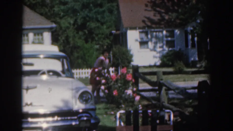 1963 New Jersey home-movie frame showing the woman tending flowers near a white car, with a house, lawn, and pink blossoms visible.