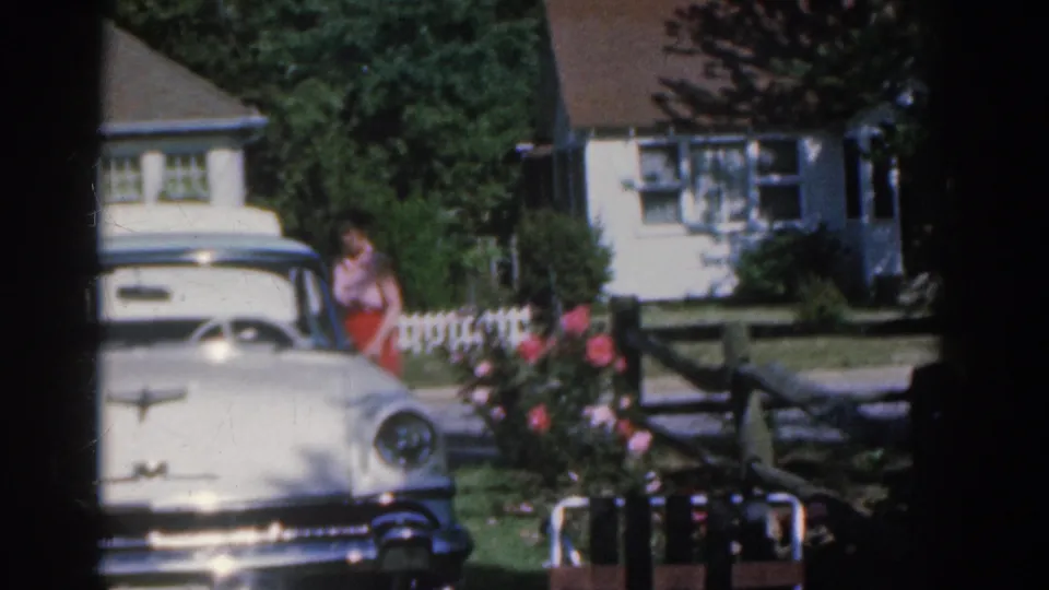 1963 New Jersey frame showing a woman near garden flowers beside a white vintage car, with a house, trees, and fence in the background.