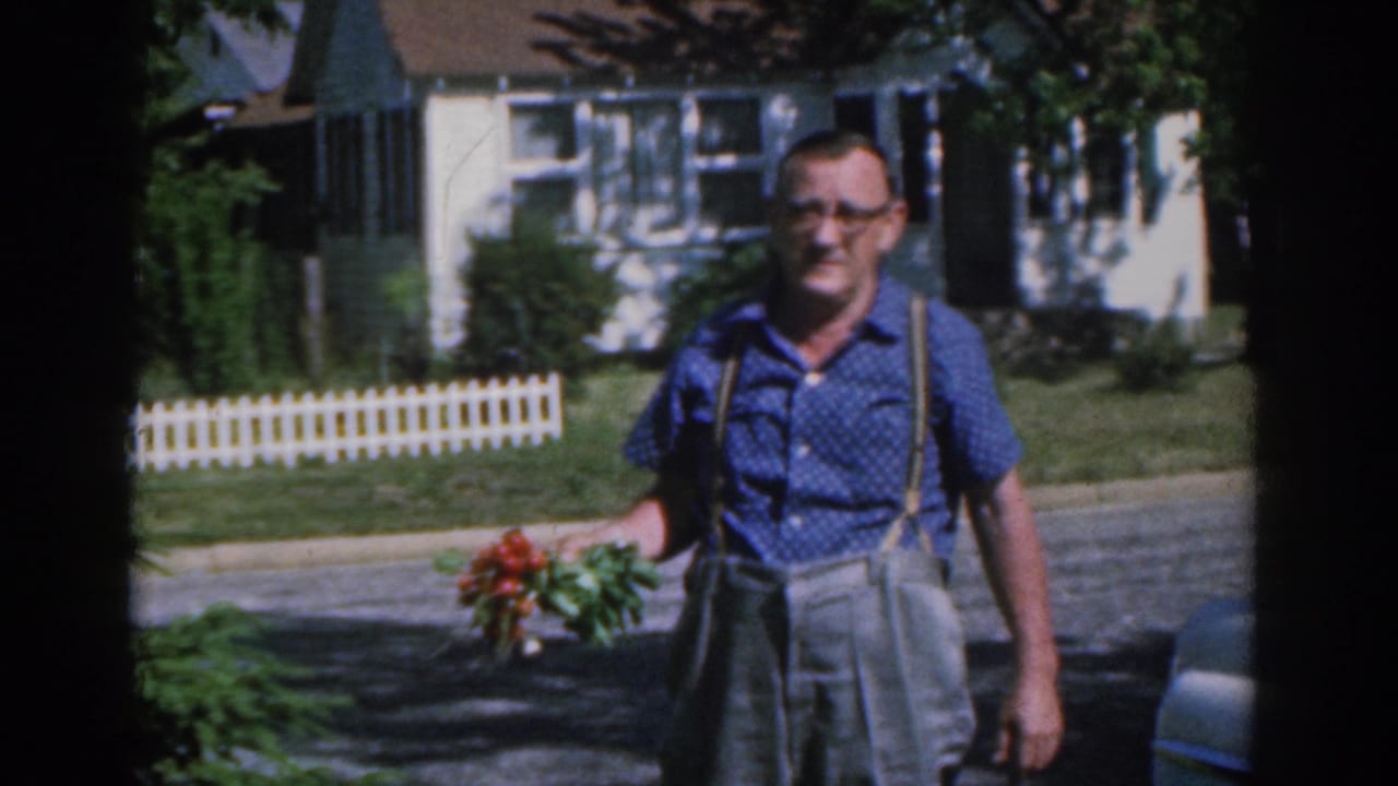In 1963 New Jersey home-movie footage, an adult man in a blue shirt and suspenders carries red roses near lawns and a white fence.