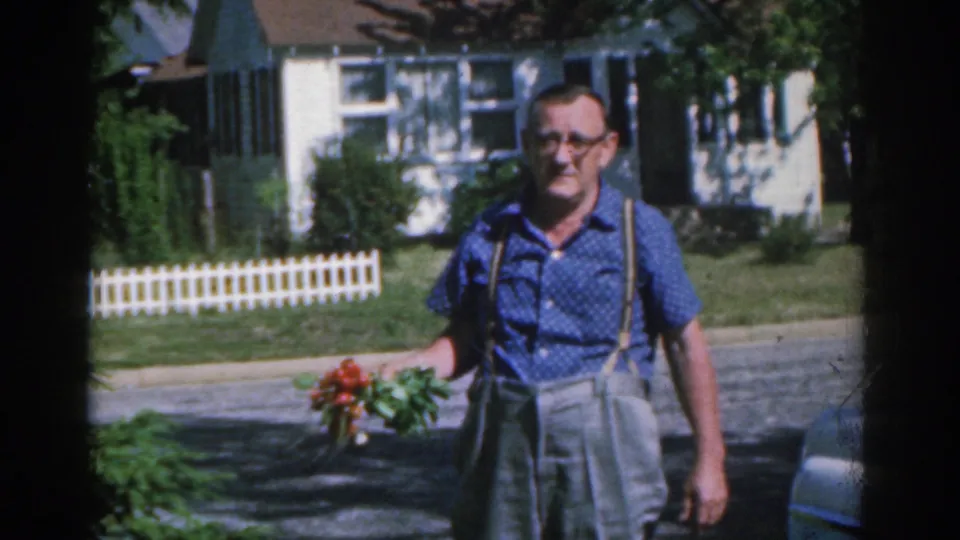 The man holding red roses is framed closer against a New Jersey suburban house, lawn, and white picket fence in 1963 8mm footage.