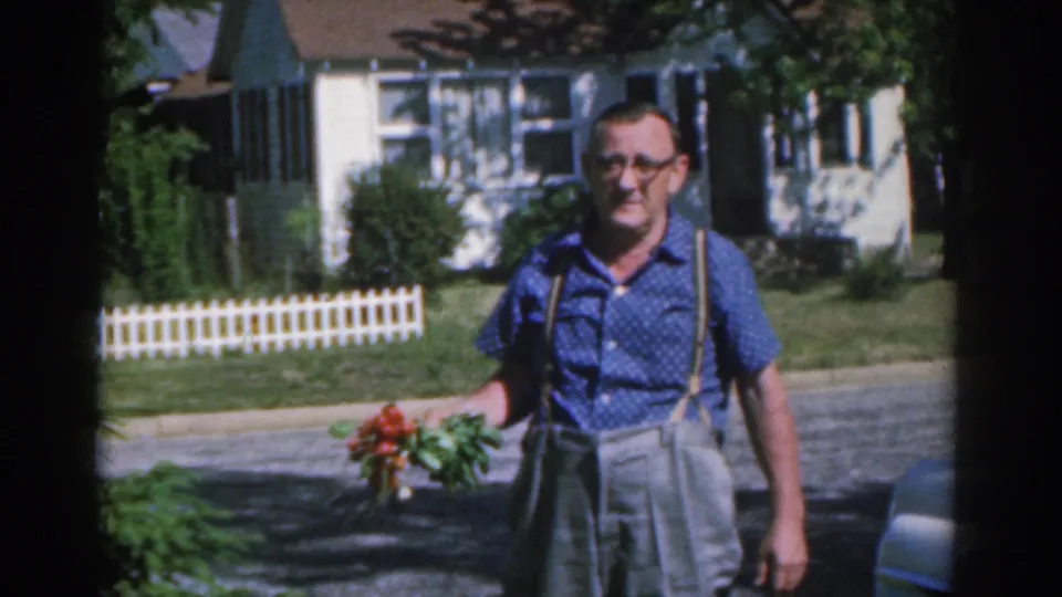 The adult man with red roses faces the camera in 1963 New Jersey footage, with a sunlit lawn, fence, and house behind him.