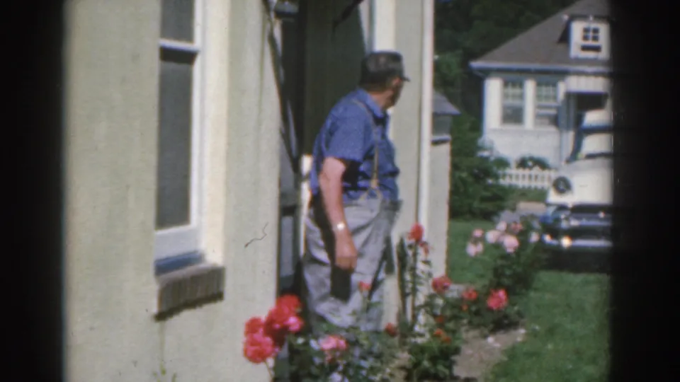 The man turns alongside a residential exterior with roses and parked cars visible, continuing the 1963 New Jersey home-movie scene.