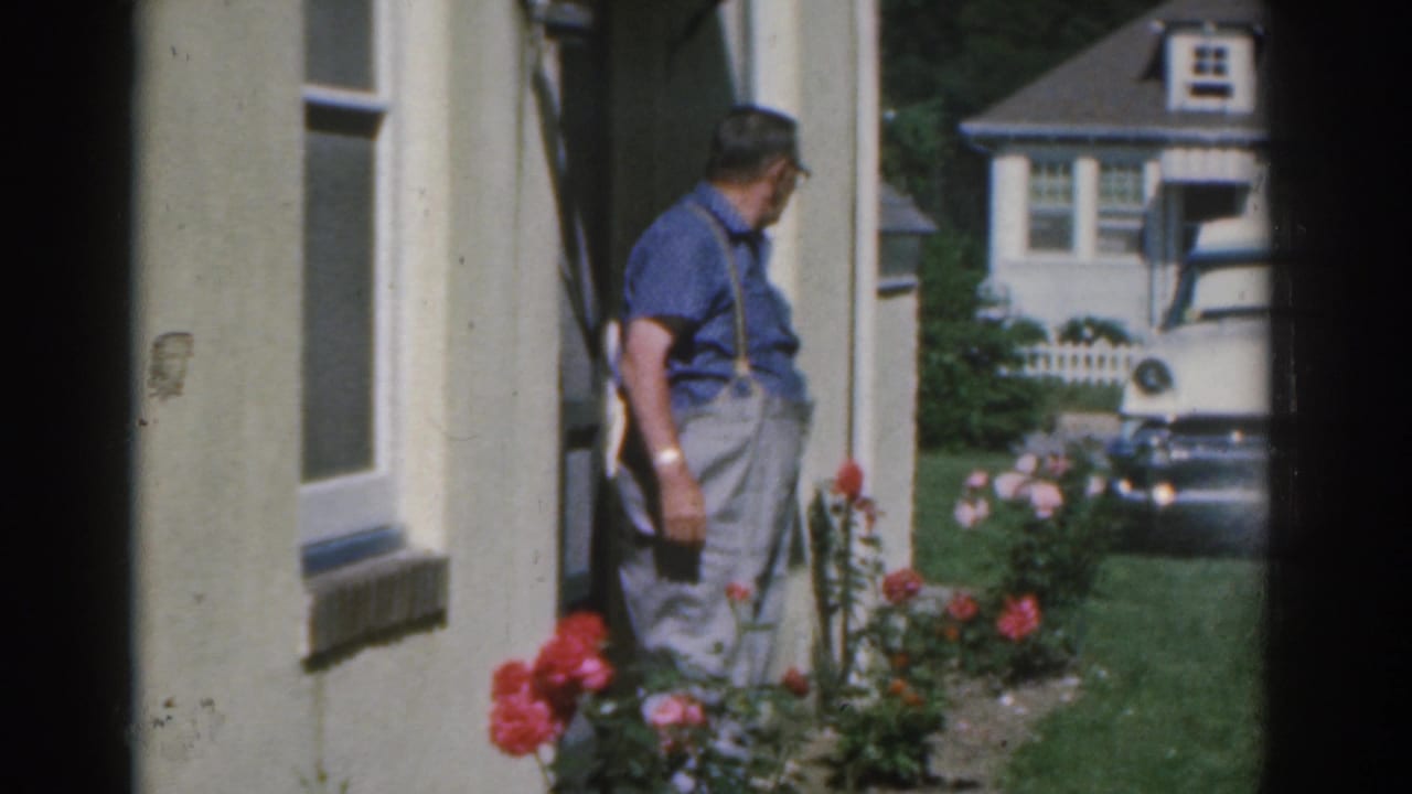 Man in blue shirt standing beside red roses near a house exterior.