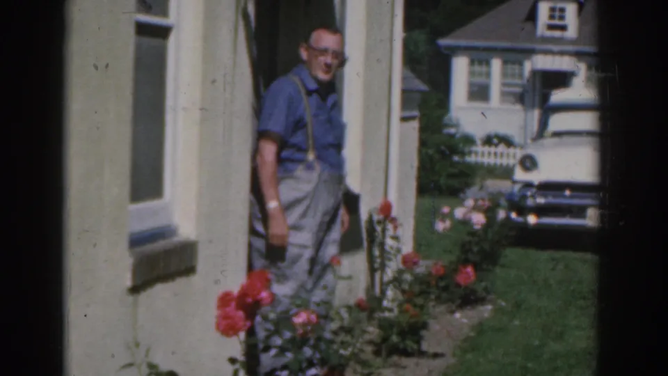 A man stands near garden roses outside a New Jersey home, with a white early-1960s car visible behind him.