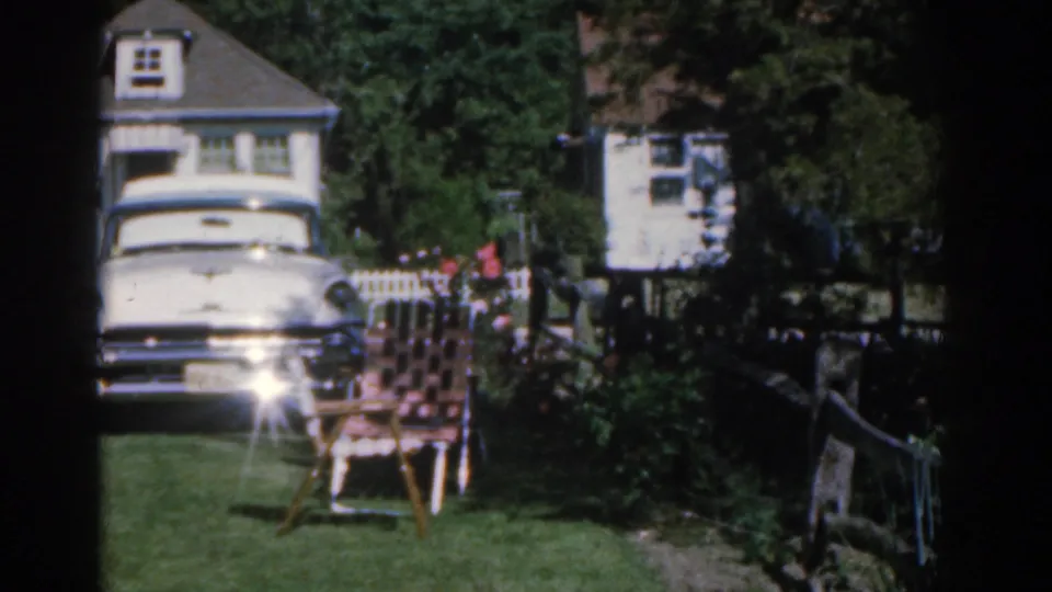 A white period car sits near a lawn and garden chair outside a suburban New Jersey house in 1963 footage.