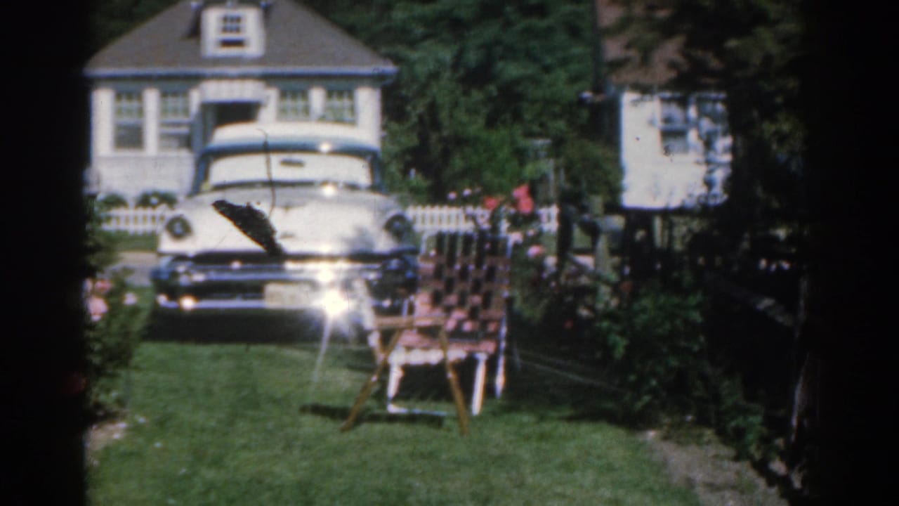 1963 New Jersey home-movie frame showing a white mid-century sedan outside a suburban house, with a green lawn, rose bushes, and a patterned lawn chair in the foreground.