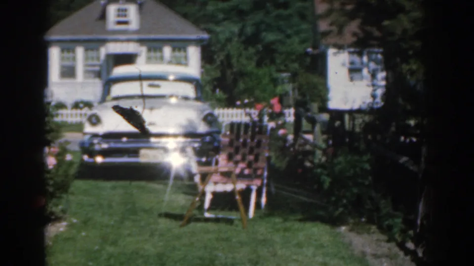 1963 New Jersey home-movie frame showing a white mid-century sedan outside a suburban house, with a green lawn, rose bushes, and a patterned lawn chair in the foreground.