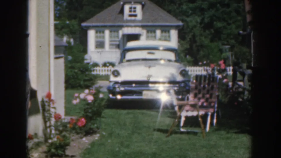 1963 New Jersey home-movie frame showing a suburban house exterior, white sedan, porch column, rose bushes, green lawn, and patterned lawn chair.