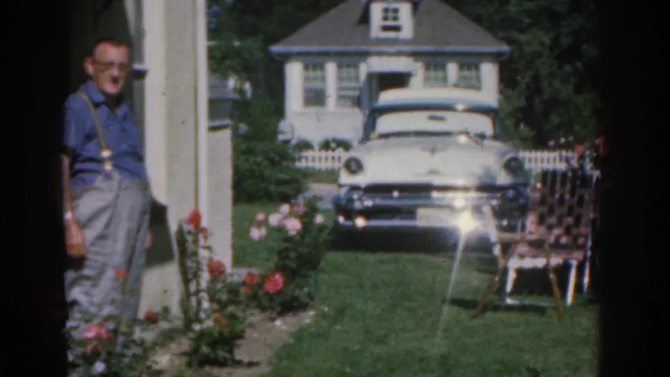 1963 New Jersey home-movie frame showing an adult man in a blue shirt and slacks standing near the porch, with a white sedan, rose bushes, lawn chair, and suburban house behind him.