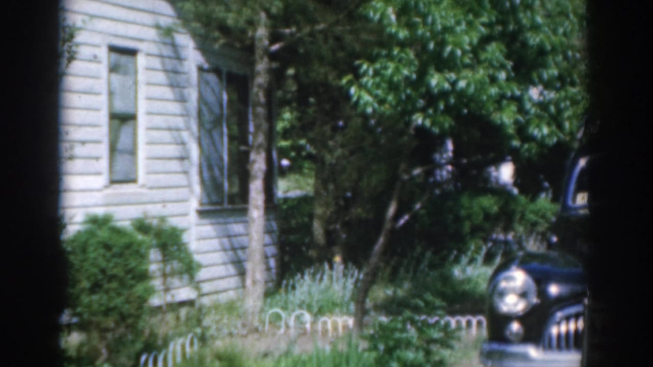 1963 New Jersey 8mm frame with a black classic car entering view near a white-sided house, shrubs, and a low fence.
