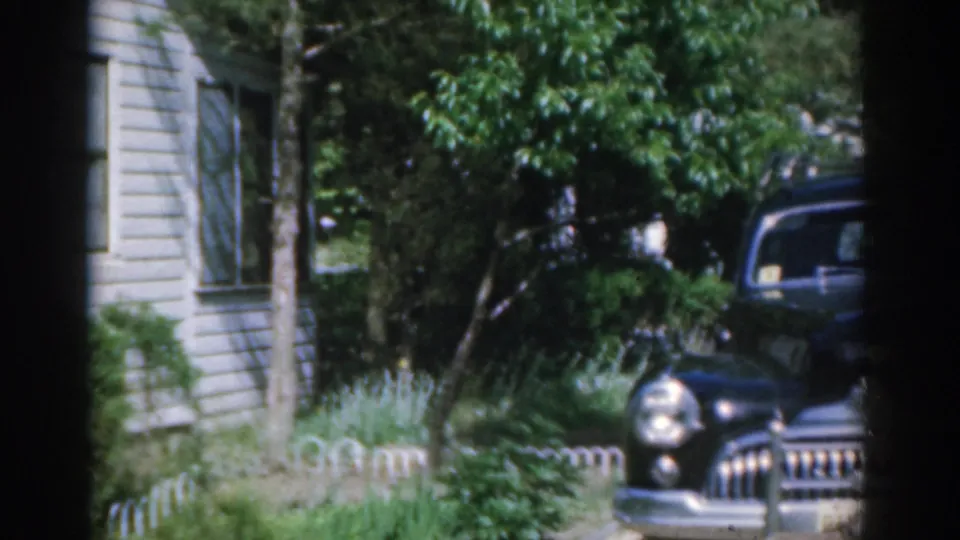 The pan shows more of the black car's chrome grille and rounded front near a white New Jersey house in 1963 footage.