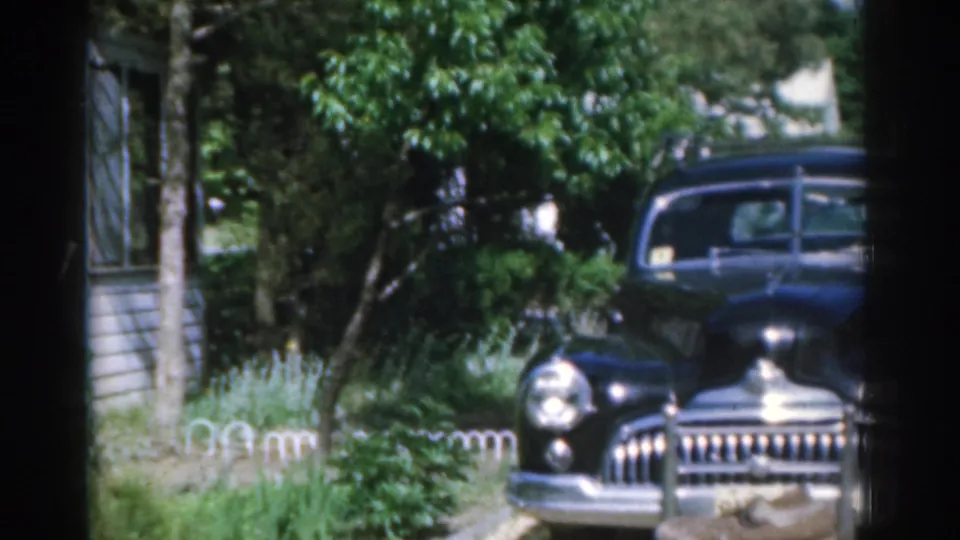 A black mid-century car sits in a suburban driveway, with chrome trim, trees, grass, and house siding visible.