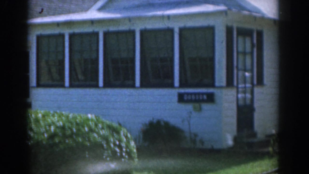 White suburban house with dark shutters, front window, lawn, hedges, and a Dobson sign.