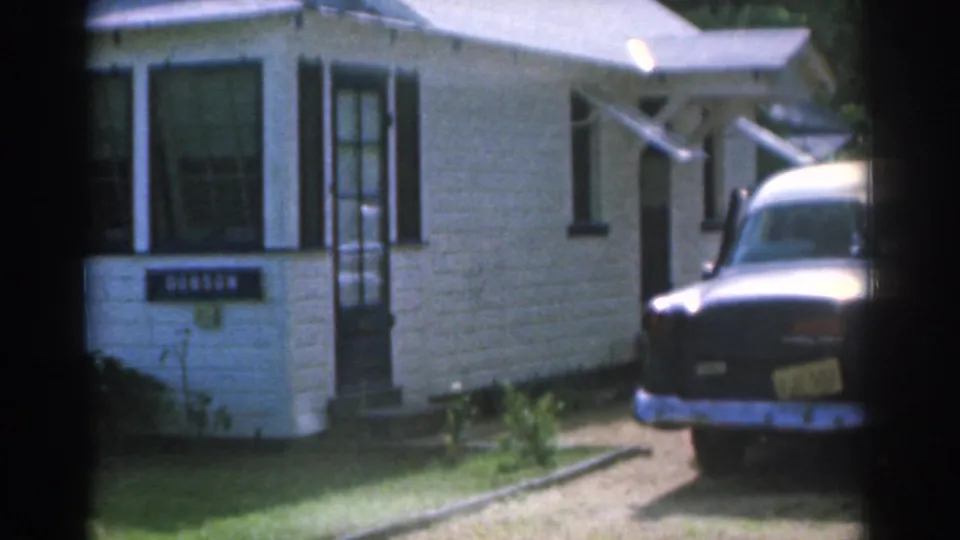 1963 New Jersey home-movie frame showing the white house, front entry, readable Dobson sign, driveway, and older sedan parked at the right.