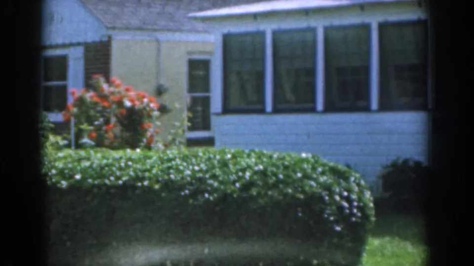 1963 New Jersey home-movie frame showing the white house from another angle with front windows, hedges, lawn, and a red flowering bush.