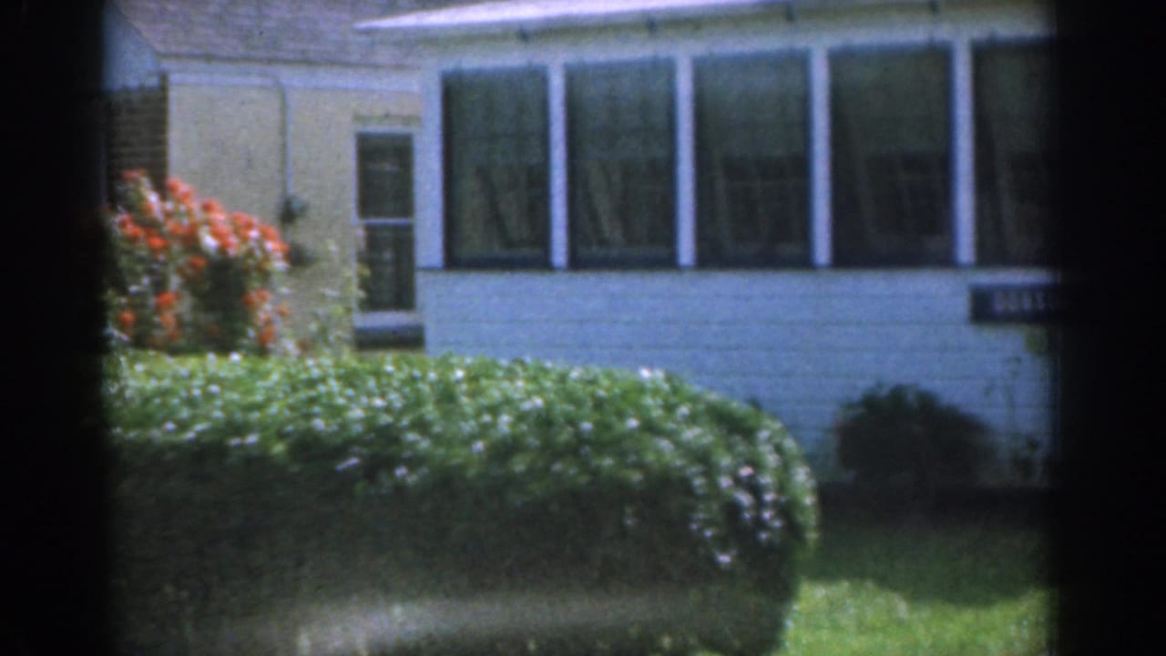 1963 New Jersey 8mm frame showing a white suburban house with sunroom windows, a green hedge, red flowers, and lawn in daylight.