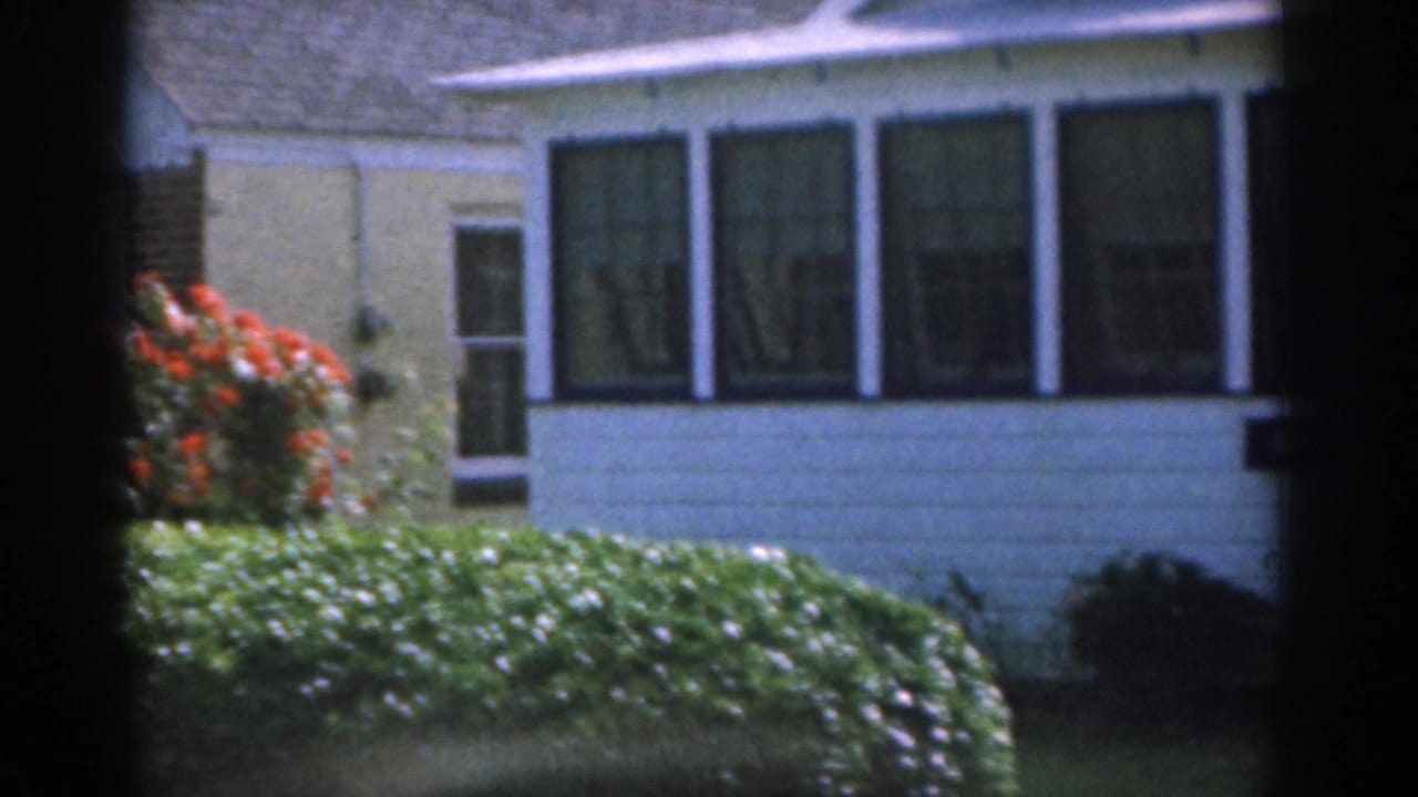 White house exterior with sunroom windows, hedge, flowers, and lawn.