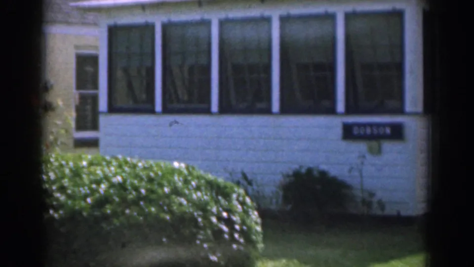 1963 New Jersey 8mm frame showing the white house sunroom, clipped hedge, lawn, and a readable nameplate near the entrance area.