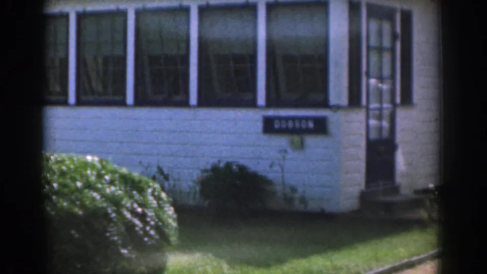 1963 New Jersey home-movie frame showing the white house facade, sunroom windows, readable nameplate, hedge, lawn, shrub, and entrance door.