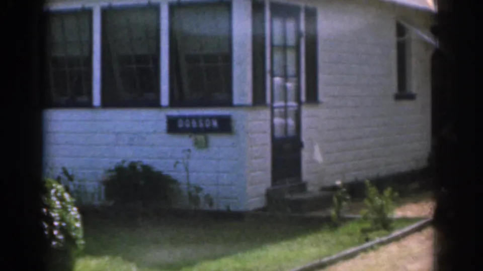 1963 New Jersey home-movie frame of a white house facade with black trim, entry steps, DOBSON sign, and sunny lawn.