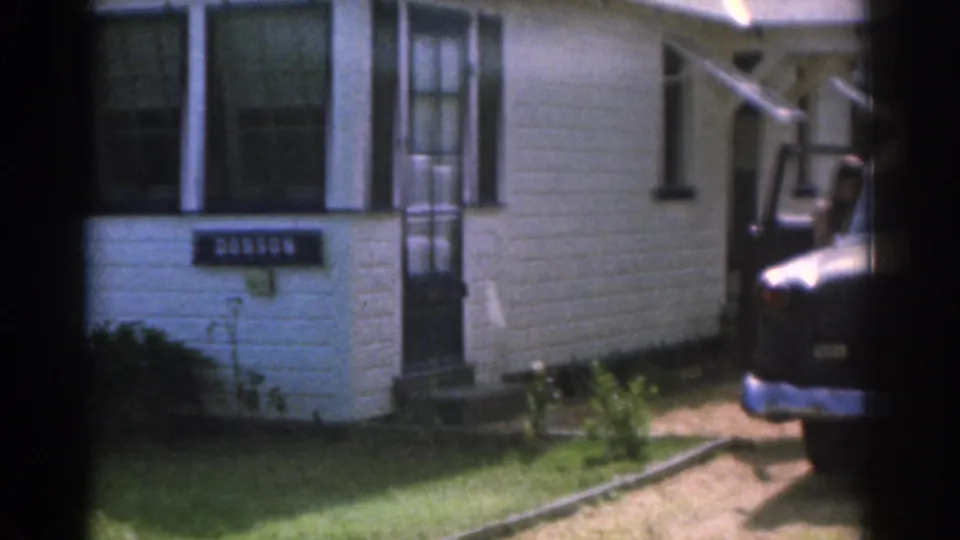1963 New Jersey 8mm view of the white house front, DOBSON sign, entry steps, lawn, and a dark car partly visible nearby.