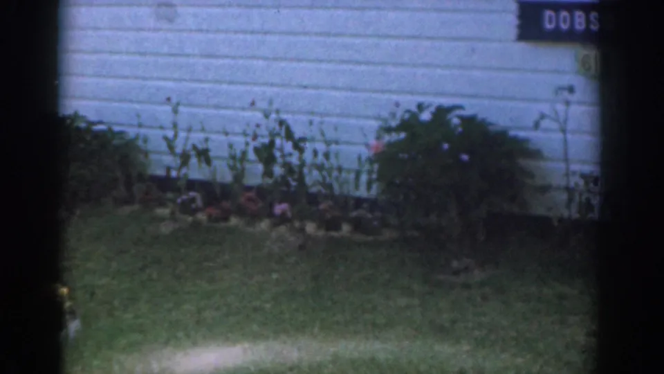 White clapboard house exterior with a DOBSON sign visible above garden plants in a 1963 New Jersey home movie.