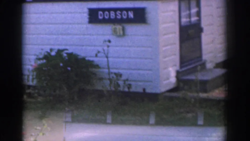 1963 New Jersey 8mm frame showing a white clapboard house exterior with a visible DOBSON sign, grass, shrubs, and part of a window.