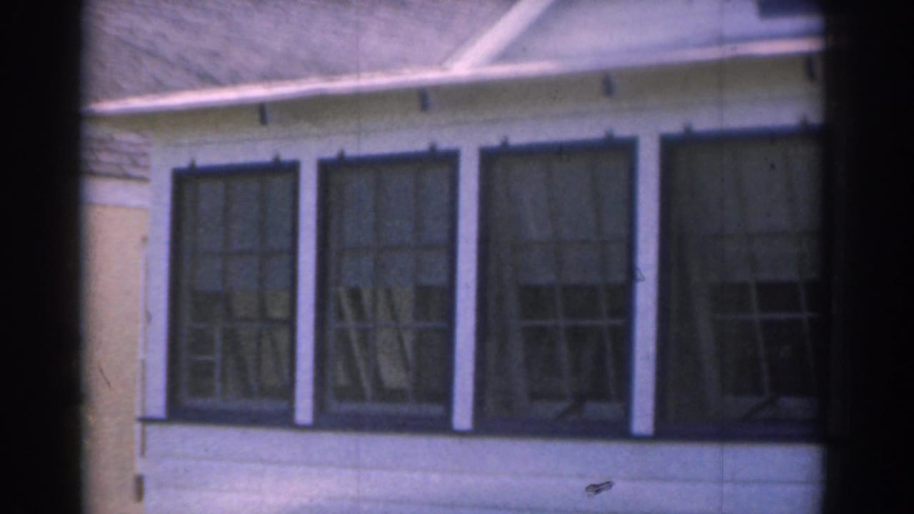 1963 New Jersey 8mm frame showing large dark-framed glass windows on a light-colored residential exterior with a sloped roof edge above.