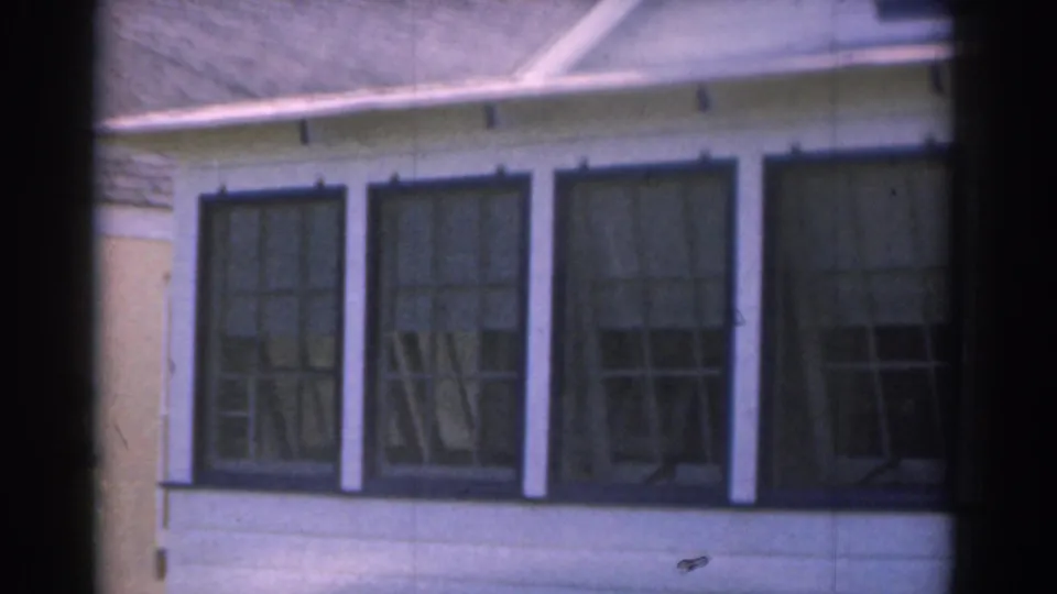 1963 New Jersey 8mm frame showing large dark-framed glass windows on a light-colored residential exterior with a sloped roof edge above.