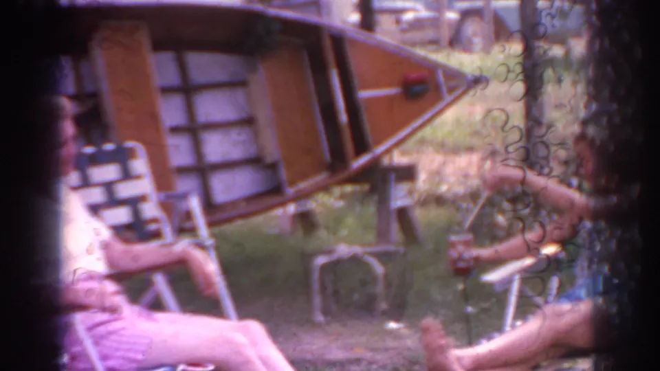 1963 New Jersey 8mm frame showing a yard with folding lawn chairs, a wooden boat on supports, grass, and partially visible seated people near the foreground.