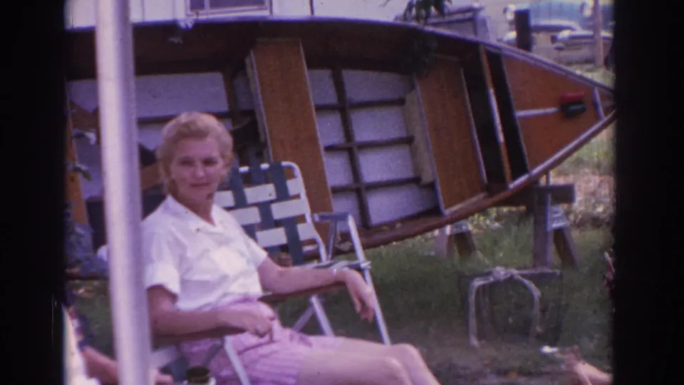 1963 New Jersey 8mm frame showing a woman in a white shirt and pink shorts seated in a folding lawn chair beside a wooden boat in a residential yard.